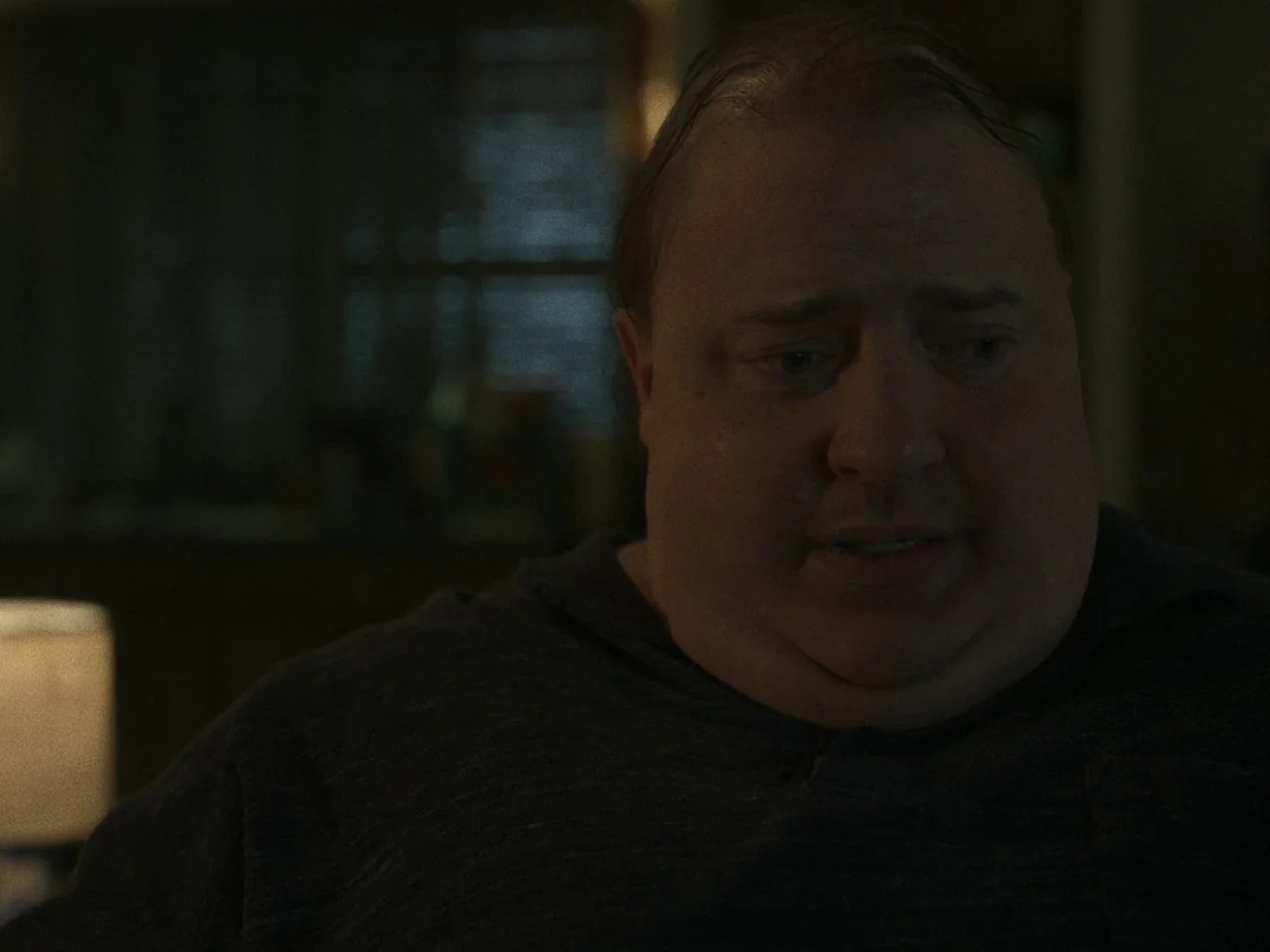The Whale (2022) movie still: closeup — A dimly lit room with a muted, dark color palette and soft shadows. A man with a heavy build sits in low…