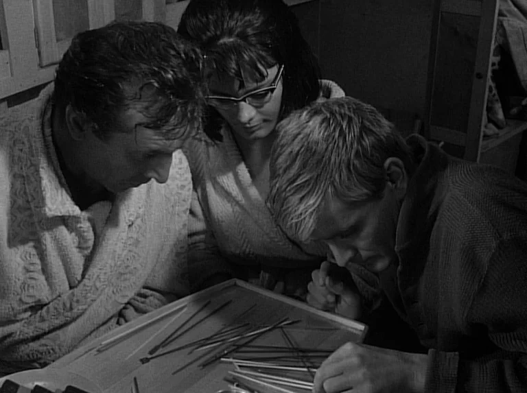 Knife In The Water (1962) movie still: medium — Three individuals lean over a shallow tray filled with various thin sticks. They observe the items…