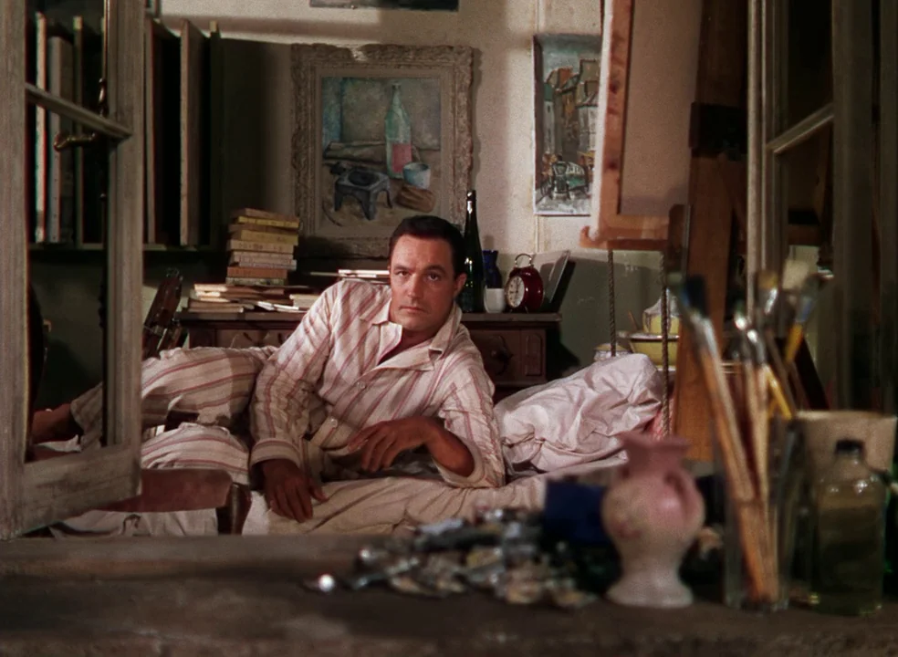 An American In Paris (1951) movie still: medium — A cluttered artist studio features framed paintings, brushes in a jar, and stacks of books. A man…