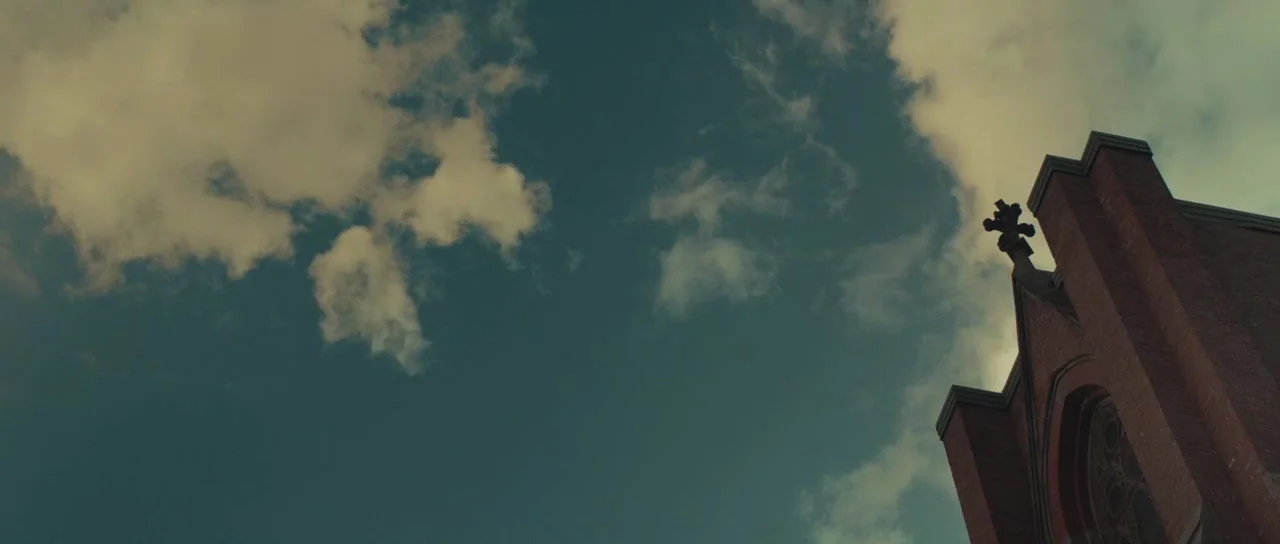 Meadowland (2015) movie still: establishing — A brick church tower with a small cross on top rises against a clouded sky. The building occupies the…