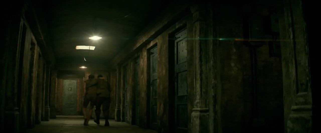 Overlord (2018) movie still: wide — A long, dark concrete corridor is lined with heavy, metal-plated cell doors. Two men in tattered uniforms walk…