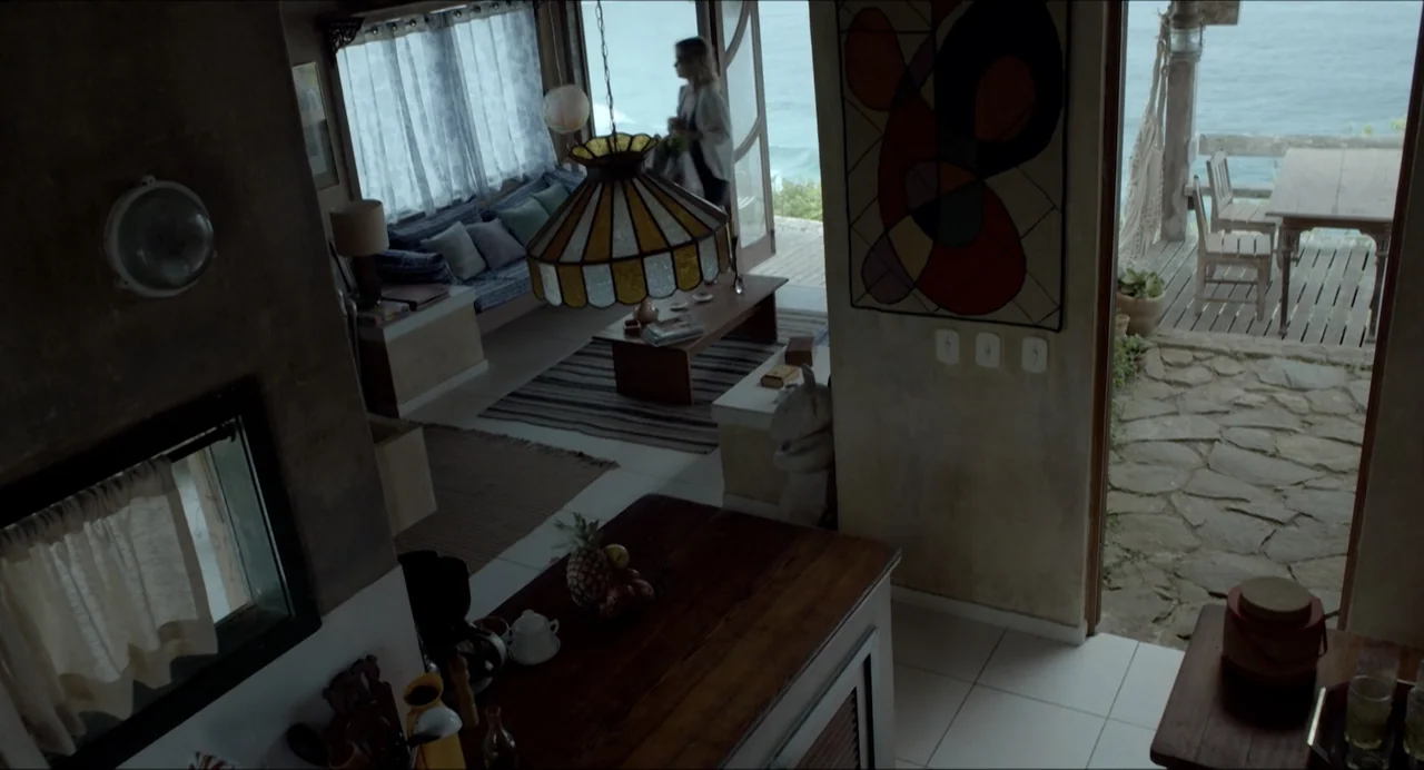 Suspension Points (2022) movie still: wide — A beach house interior features a wooden kitchen counter and a living area. A woman walks inside…