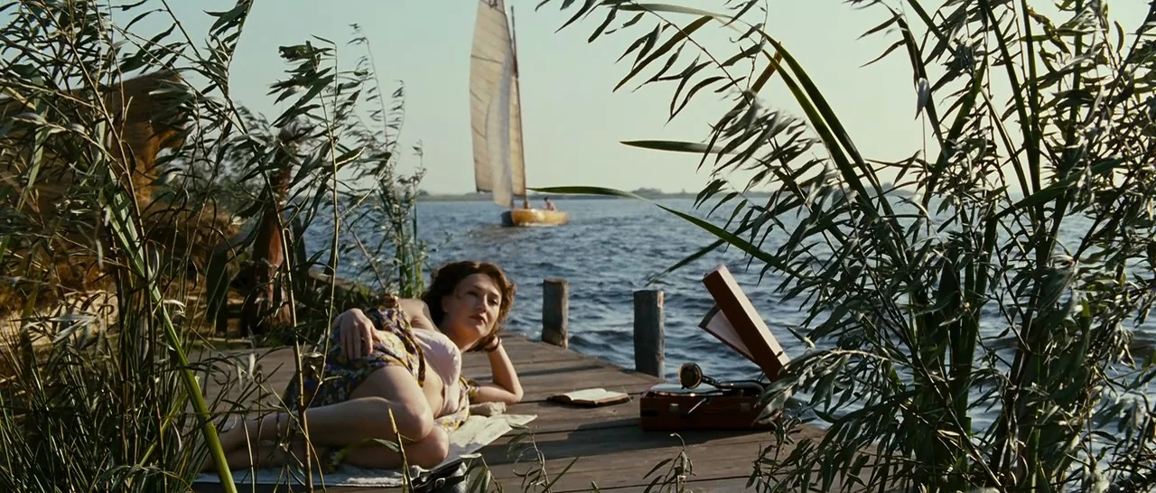 Black Book (2006) movie still: wide — A wooden pier extends into a calm lake surrounded by tall green reeds. A woman in a summer dress rests on a…