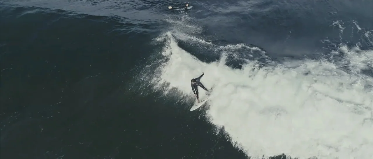 Taking Back the Waves (2017) movie still: wide — A surfer rides a wave in a vast expanse of dark ocean water. The surfer balances on a white board…