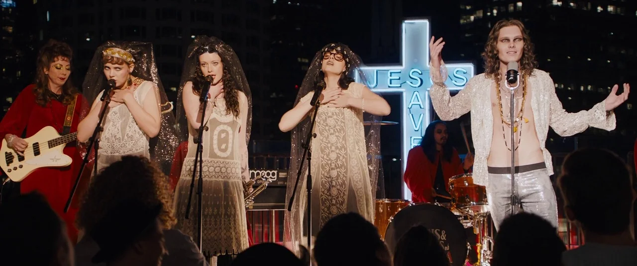 Under The Silver Lake (2018) movie still: wide — A musical performance takes place on a rooftop at night with city skyline lights. Several musicians…