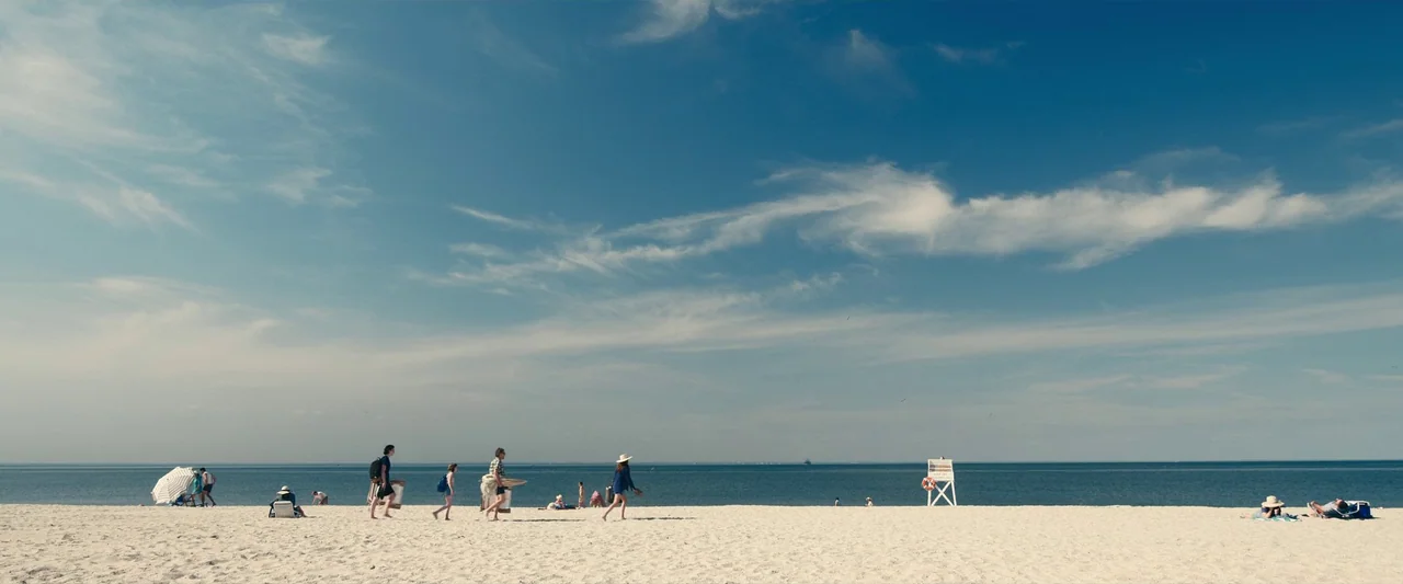 Leave The World Behind (2023) movie still: wide — A wide stretch of white sand meets the horizon under a clear blue sky. Several individuals are…