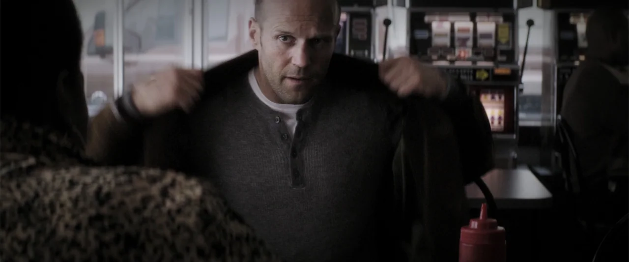 Wild Card (2015) movie still: medium — A dimly lit casino interior featuring slot machines and tables in the background. A man with a shaved head is…