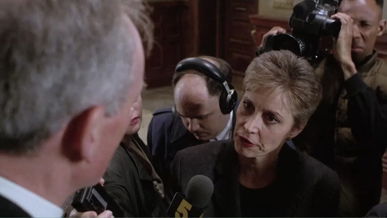 Mistrial (1996) movie still: medium — A group of reporters gathers in an interior space holding microphones and camera equipment. A woman in a dark…