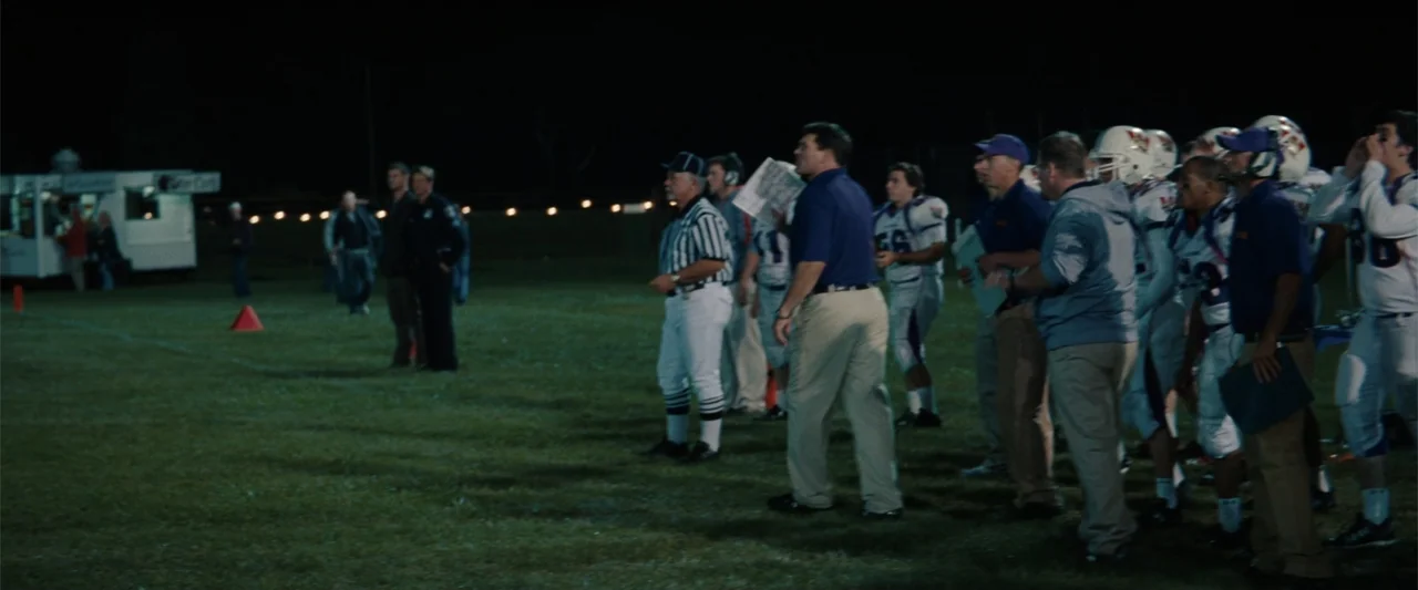 Red Dawn (2012) movie still: wide — A night time football field with coaches and players standing along the sideline grass. A group of men in…