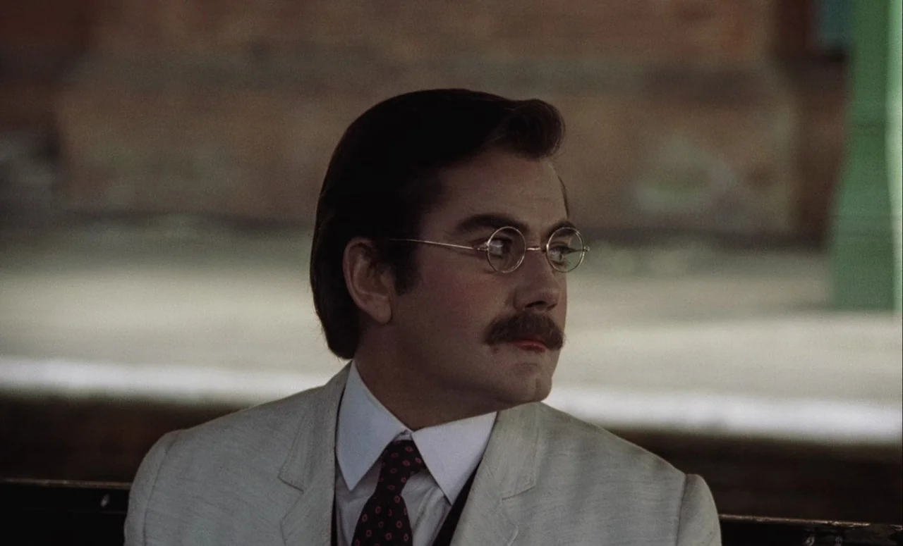 Mahler (1974) movie still: closeup — A man stands on a public transit platform with blurred structural elements behind him. He wears a formal suit…