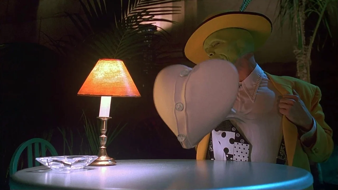The Mask (1994) movie still: medium — A dimly lit club setting features a round table with a small lamp. A man wearing a green mask sits at the…