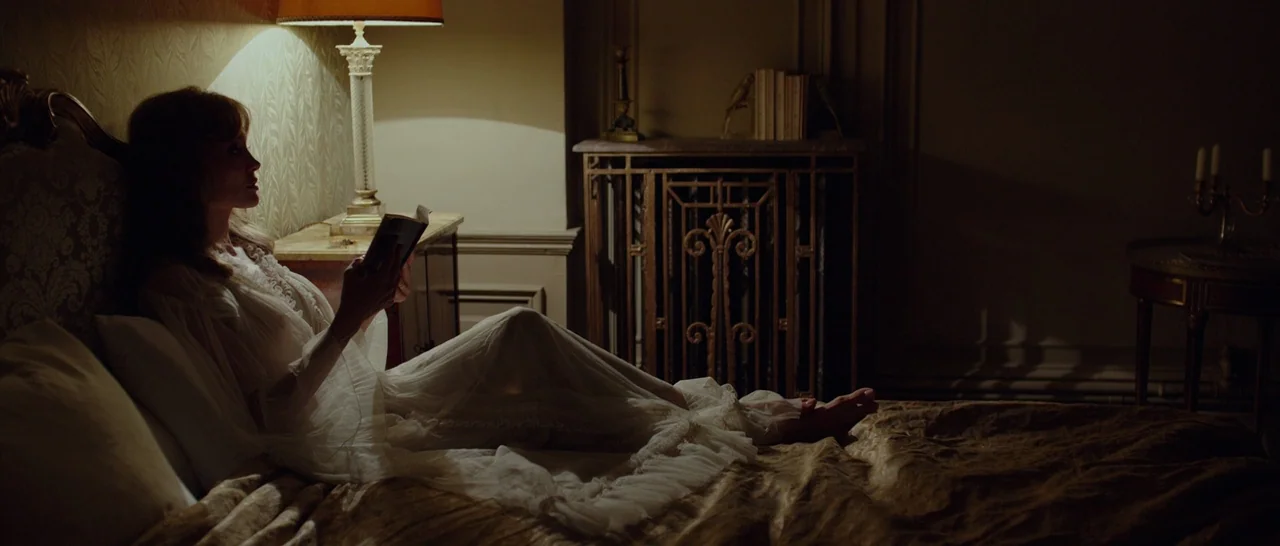 By The Sea (2015) movie still: medium — A dimly lit hotel room features an ornate metal heater and patterned wallpaper. A woman wearing a sheer…