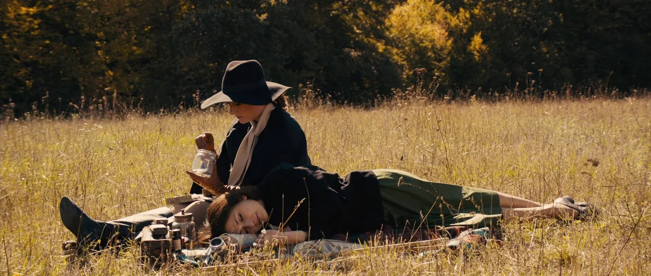 The Duke of Burgundy (2014) movie still: fullbody — An open meadow with dry, golden grass backed by a dense tree line. Two women sit on the ground…
