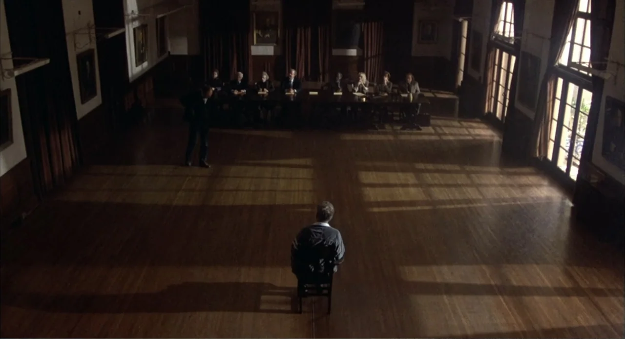Back to School (1986) movie still: wide — A spacious wood-paneled hall features tall windows on one side and a long table. A man sits alone on a…