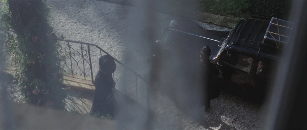 Suite Française (2015) movie still: wide — A gravel driveway with a rose-covered metal railing and a black vintage vehicle. Two people in dark coats…