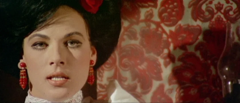Django Kill (1967) movie still: closeup — A woman stands before a wall covered in intricate red and cream damask wallpaper. She stares forward with…