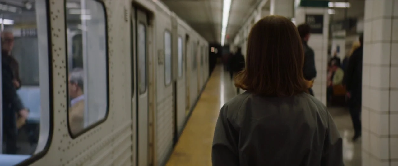 Greta movie still: medium — A woman walks away from the camera on a concrete subway platform. White tiled support columns stand near a stationary…
