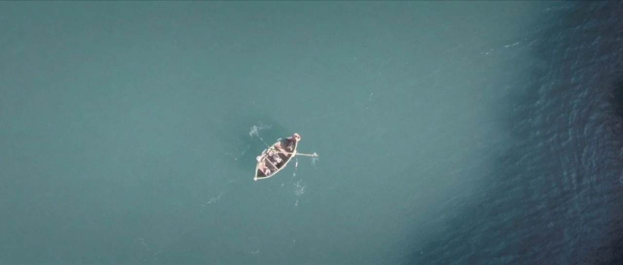 Spring (2014) movie still: establishing — A small wooden rowboat sits in the center of vast, calm, teal-colored ocean water. Several occupants row…