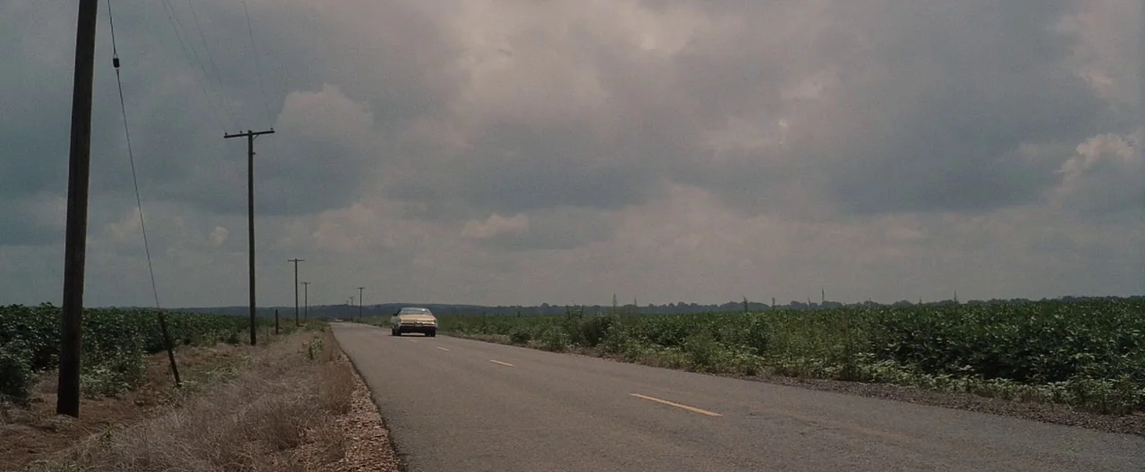 Ain’t Them Bodies Saints (2013) movie still: wide — An asphalt road stretches through a vast rural landscape bordered by green agricultural fields.…