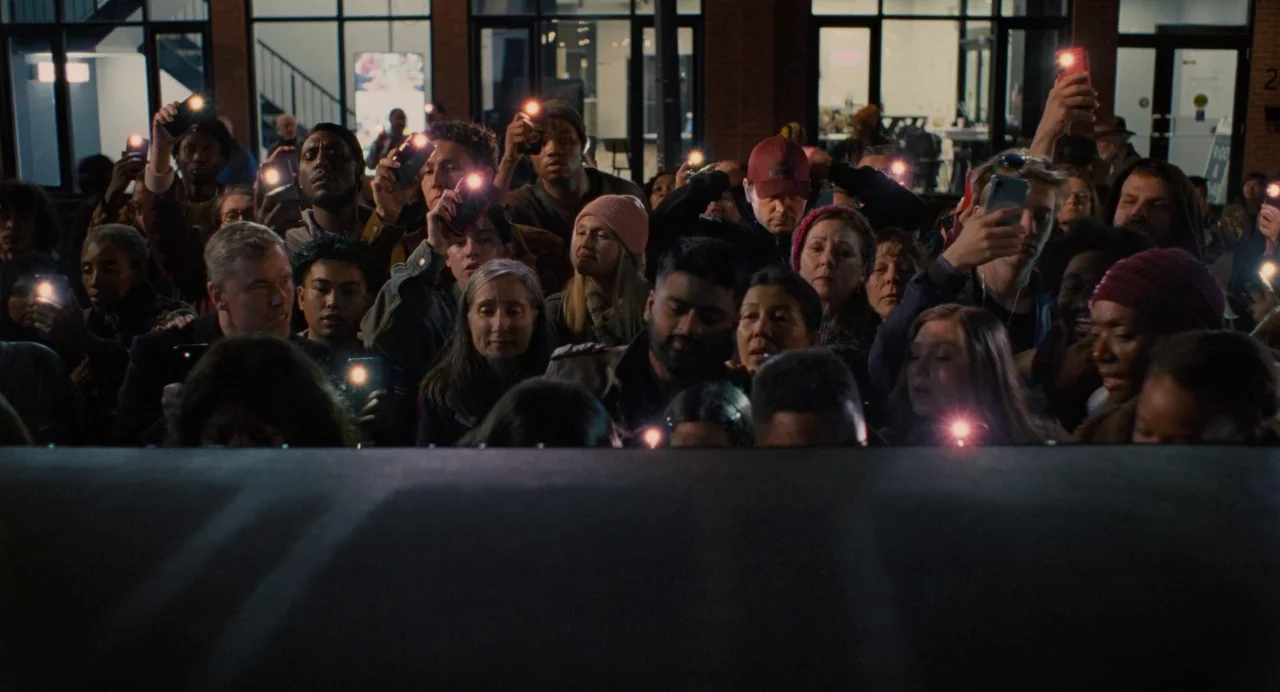 Trap (2024) movie still: medium — A diverse crowd of people stands at night in front of dark storefront windows. Many individuals hold up mobile…