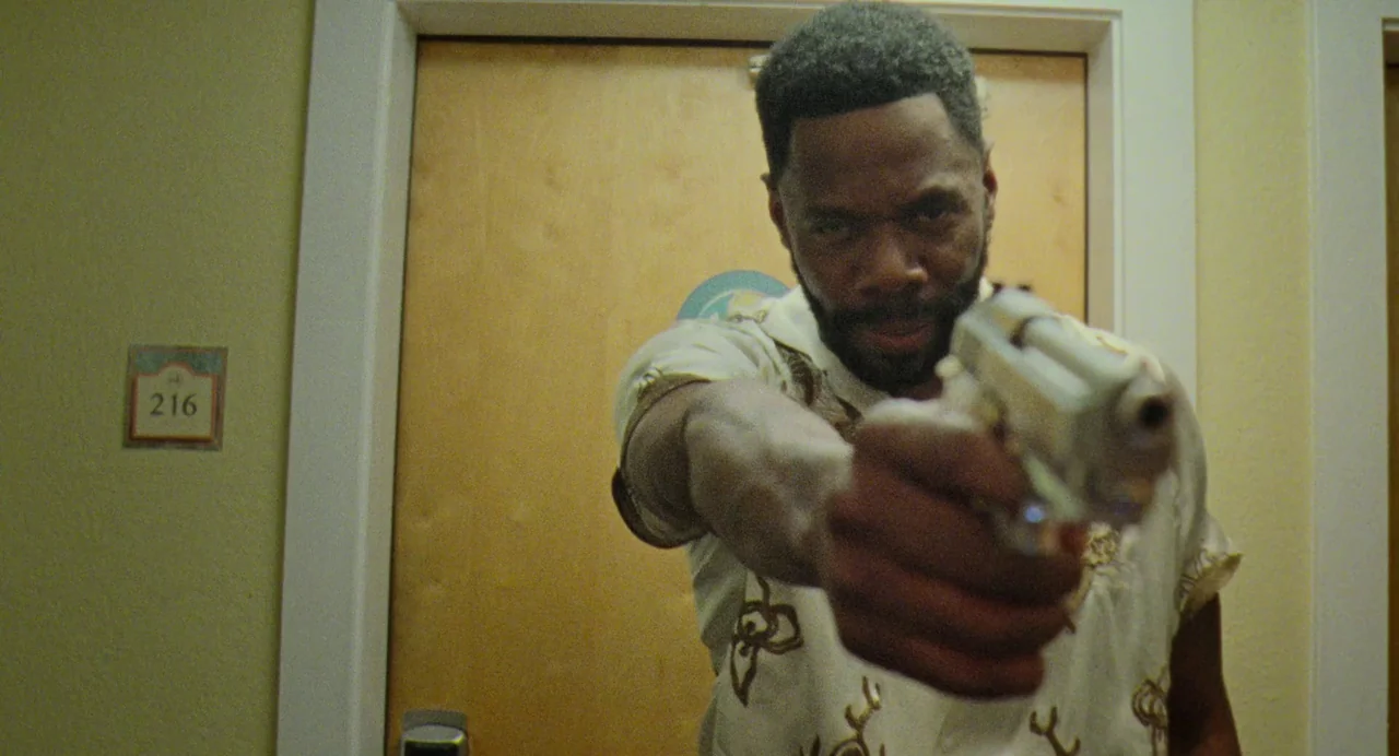 Zola (2020) movie still: medium — A hotel hallway with a door marked with the number 216. A man stands in front of the closed door aiming a small…