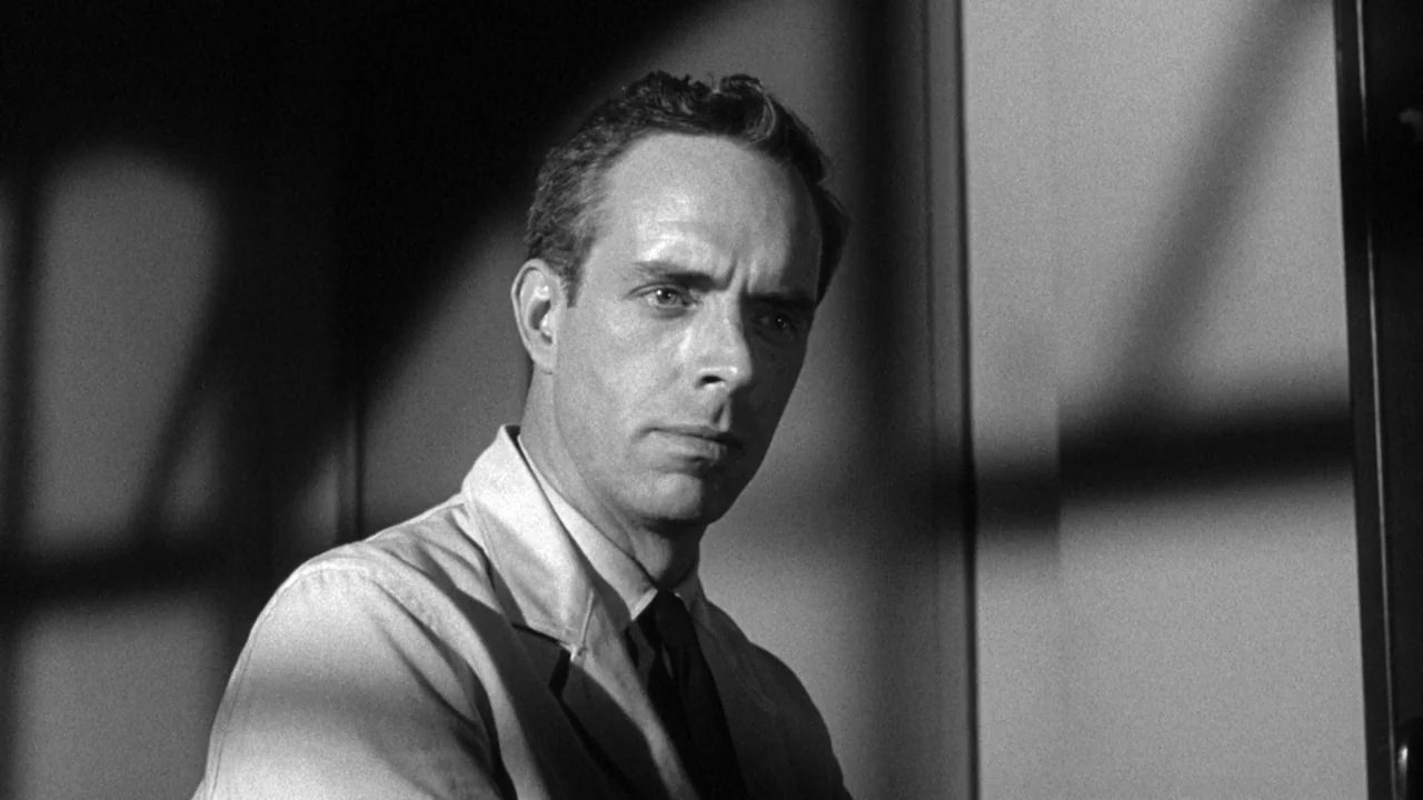 Shock Corridor (1963) movie still: medium — A dimly lit space features a man positioned against a paneled wall. He gazes off-camera while hard…