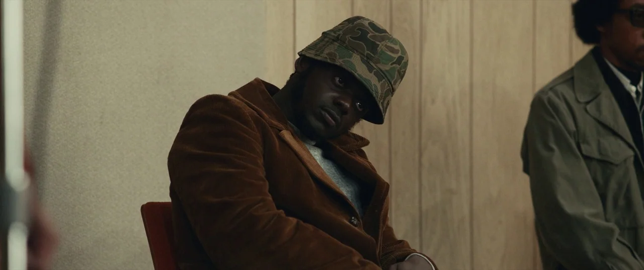 Judas and the Black Messiah (2021) movie still: medium — A room with light-colored vertical wood paneling and a plain textured wall. A man sits in a…