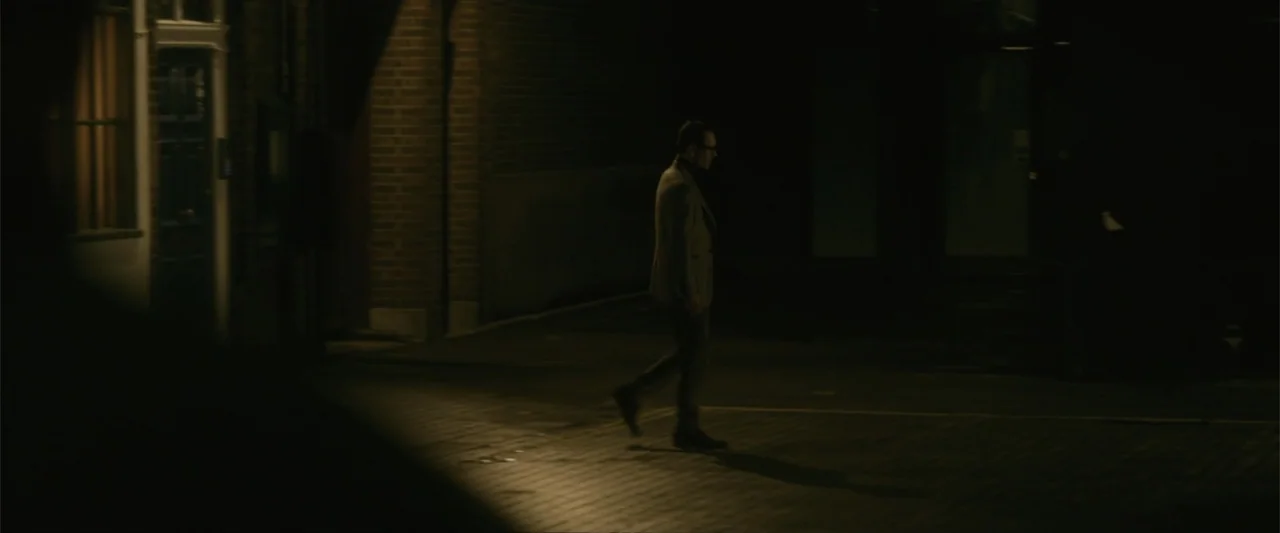 Black Bag (2025) movie still: fullbody — A dark outdoor urban space features brick architecture and a single cone of illumination. A man walks…