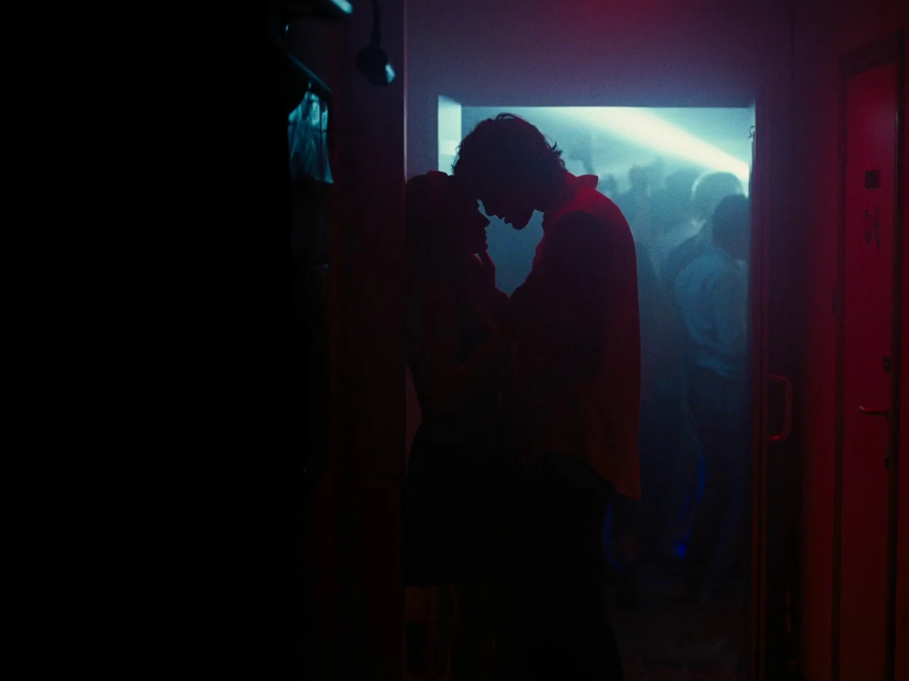 Saltburn (2023) movie still: medium — A dark, narrow doorway frames two figures standing close together against a bright club. In the background,…
