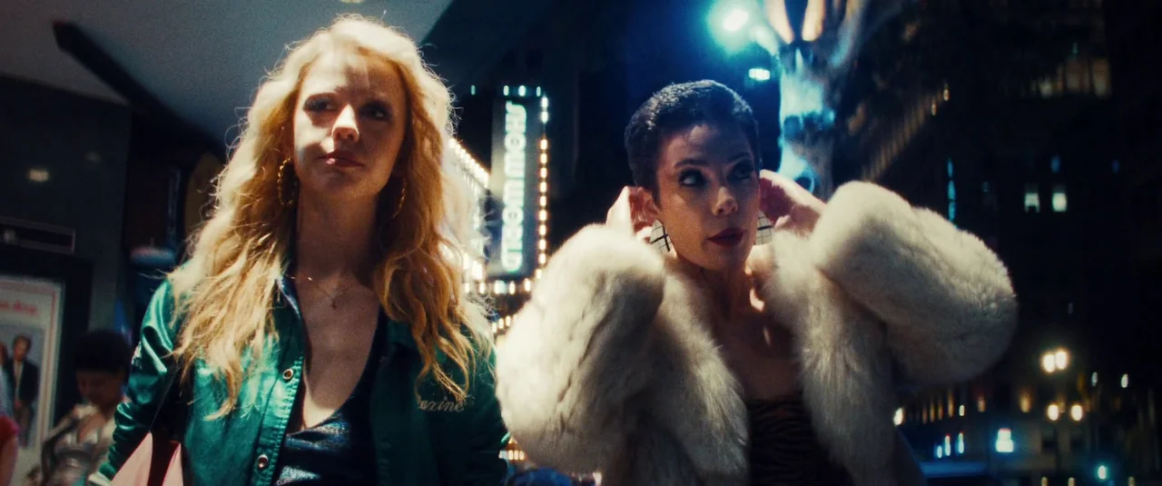 MaXXXine (2024) movie still: medium — A night scene on a city street featuring a neon Show World sign. Two young women walk past the building while…