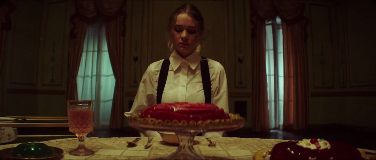 Braid (2018) movie still: medium — A formal dining room features light green walls and floor-to-ceiling draped windows. A young woman sits at a…