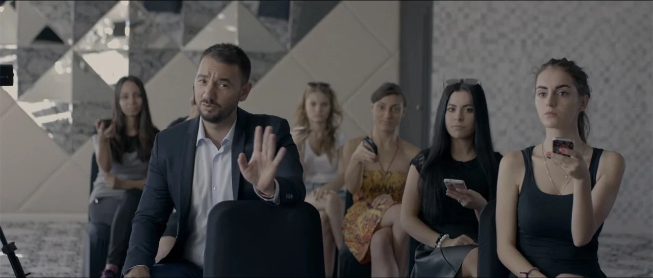 All She Wrote (2018) movie still: medium — A modern waiting room features patterned walls and multiple people sitting in chairs. A man in a blazer…