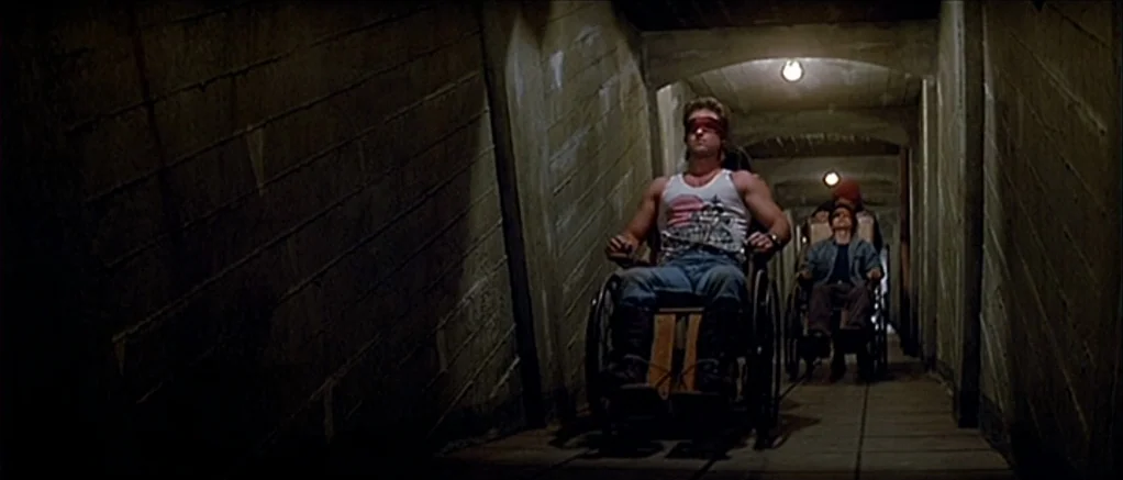 Big Trouble in Little China (1986) movie still: fullbody — A narrow concrete corridor with angled walls and dim overhead lights stretches into…