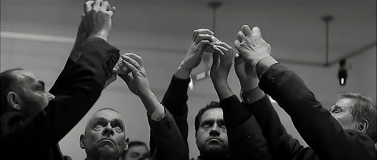 13 Tzameti (2006) movie still: medium — An interior space with plain walls and overhead lights. Several men in dark suits reach their hands upward…