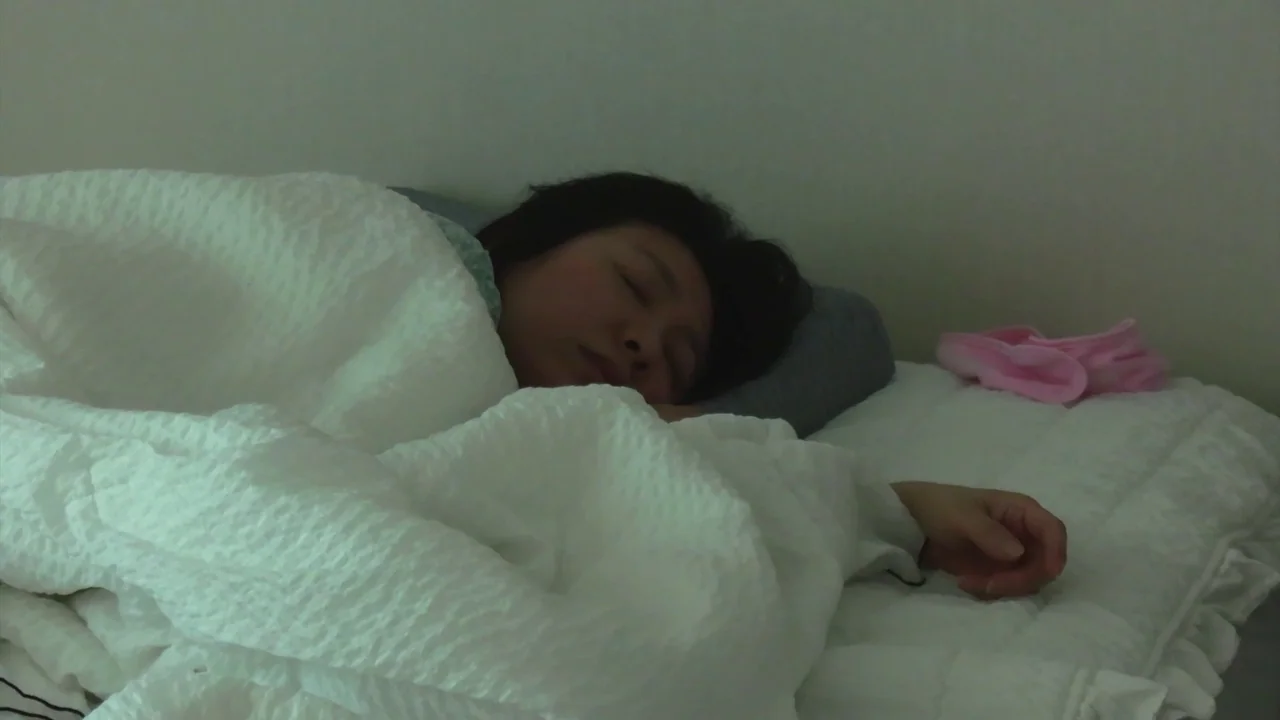 In Front Of Your Face (2021) movie still: medium — A person sleeps in a white bed against a plain, off-white wall. The subject is partially covered…