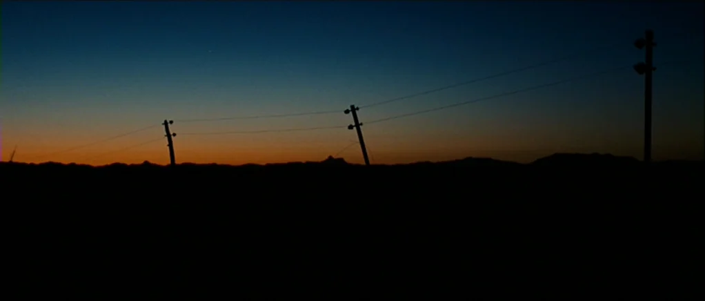 No Country For Old Men movie still: establishing — A dark, flat desert landscape stretches under a darkening sky at twilight. Utility poles and…