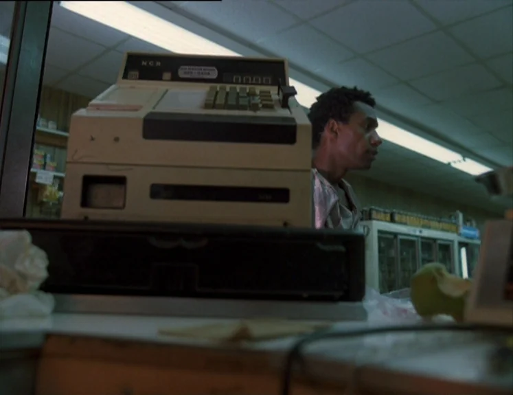 The Brother From Another Planet (1984) movie still: medium — A dimly lit retail store interior with a beige NCR cash register in the foreground. A…