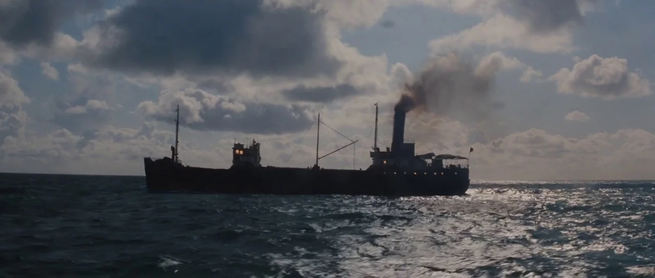 Raiders Of The Lost Ark (1981) movie still: wide — A dark cargo ship steams across the open ocean under a clouded sky. A plume of black smoke rises…