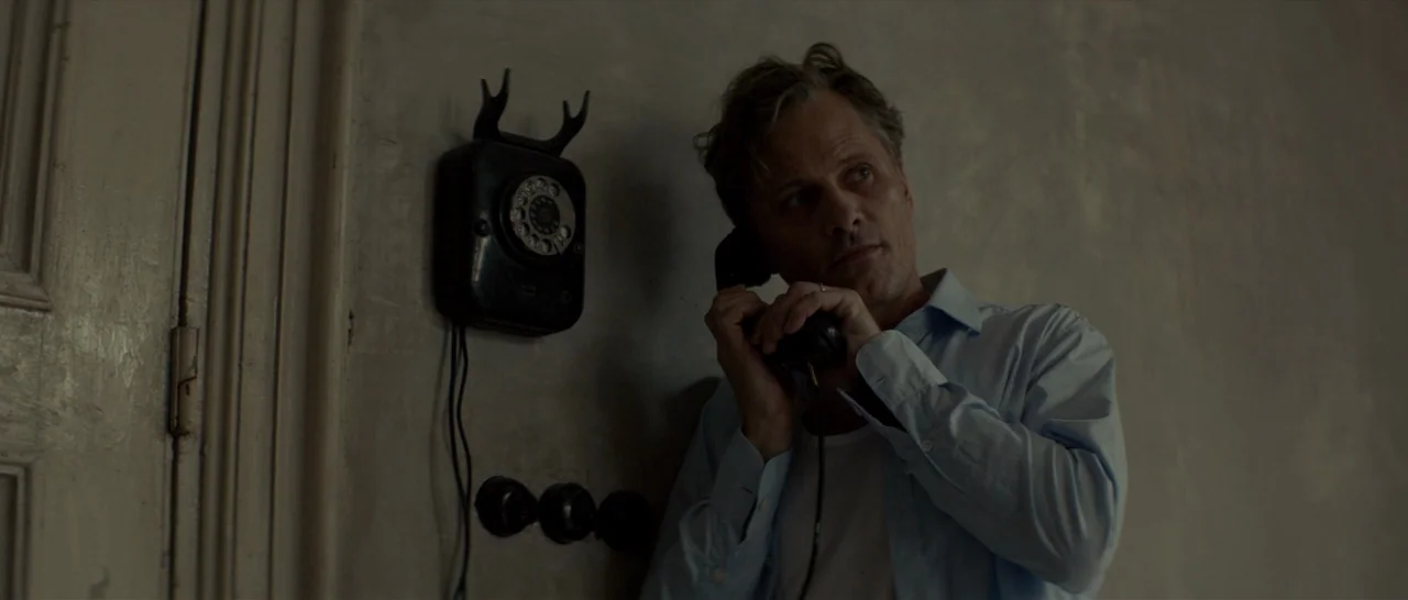 The Two Faces of January (2014) movie still: medium — An interior space features a weathered plaster wall with a vintage mounted telephone. A man in…