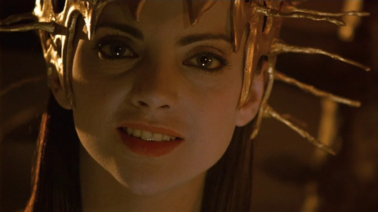 Dagon (2001) movie still: closeup — A dimly lit stone environment with dark, textured backgrounds. A woman wearing a gold spiked headpiece looks…