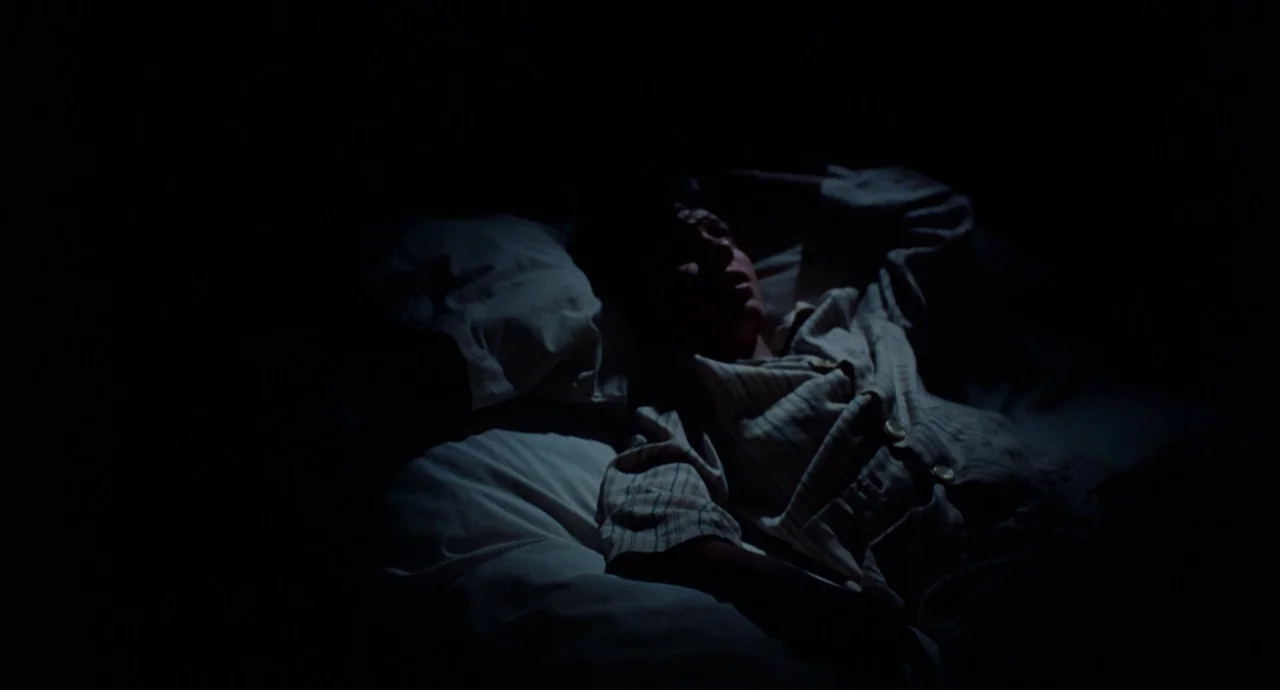 The Devils Backbone (2001) movie still: medium — A dark, dimly lit room contains a person lying on white sheets. The individual is positioned on a…