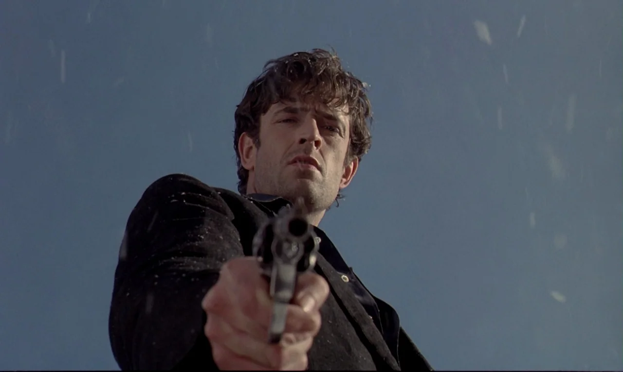 Dellamorte Dellamore (1994) movie still: medium — An outdoor location with a clear blue sky and visible dust particles. A man in a dark jacket aims…