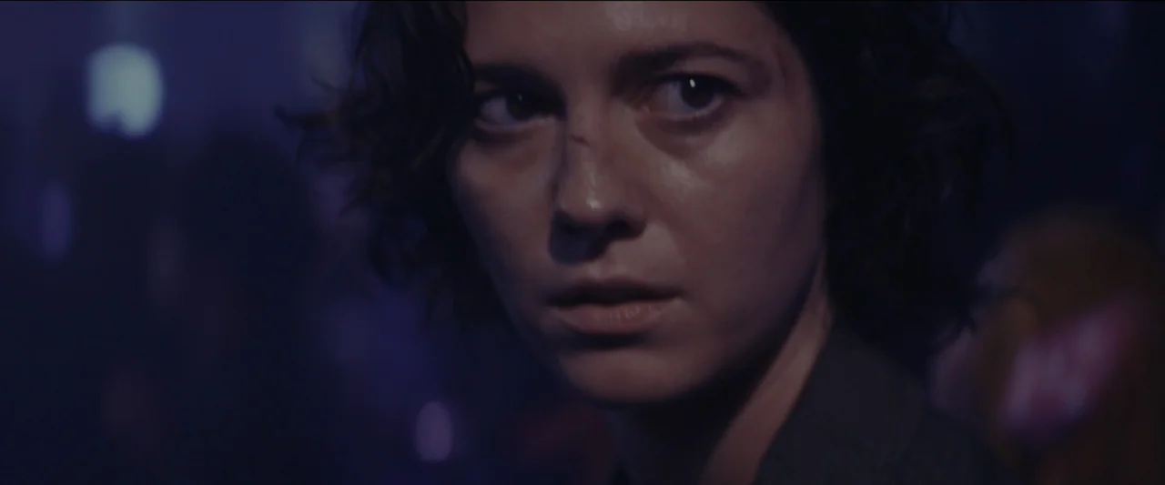 Kate (2021) movie still: closeup — A dimly lit, purple-hued space filled with ambient light suggesting a crowded venue. A woman with dark wavy hair…