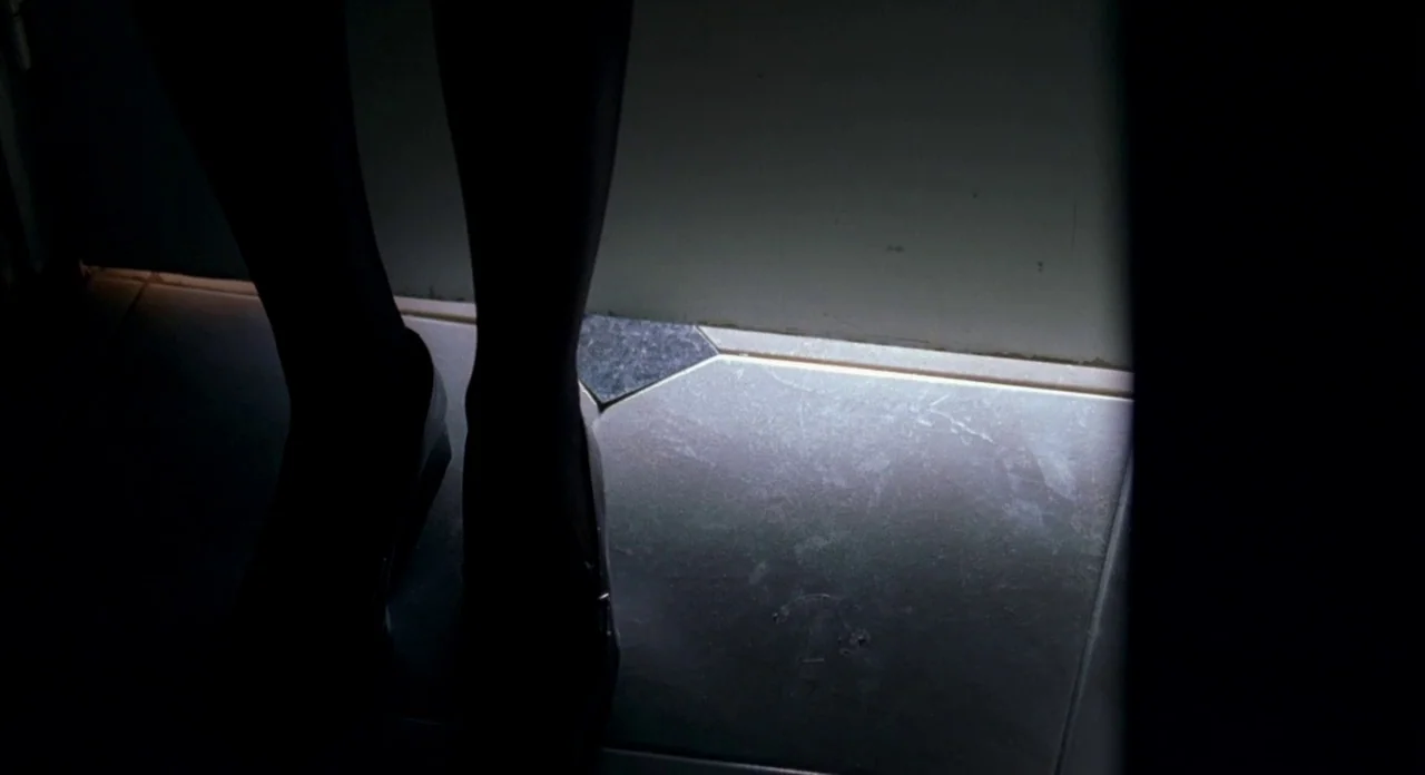 Read My Lips (2001) movie still: closeup — A dimly lit indoor hallway with a tiled floor and a closed doorway. A person stands facing a shut door…