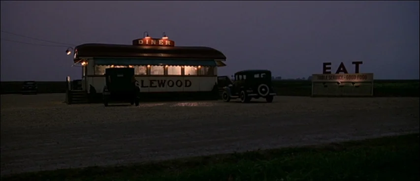 Road To Perdition (2002) movie still: wide — A remote diner sits in a flat, dark field at dusk. Vintage cars are parked on the gravel lot next to an…