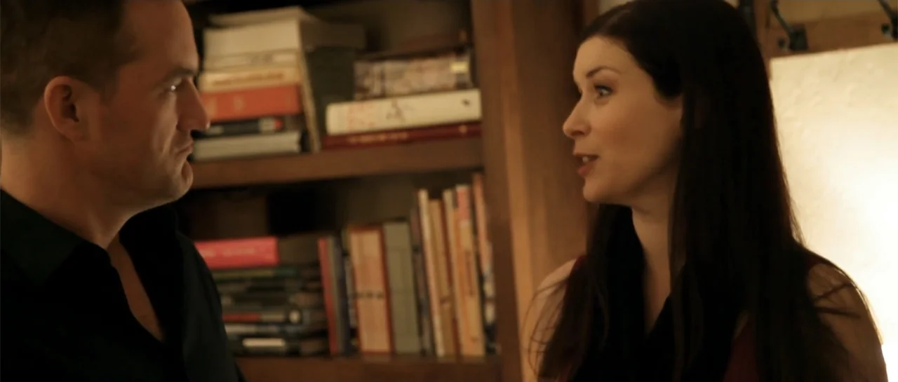 La coherencia del caos movie still: medium — A wooden library shelf with numerous books sits between two individuals. A man and a woman stand close…