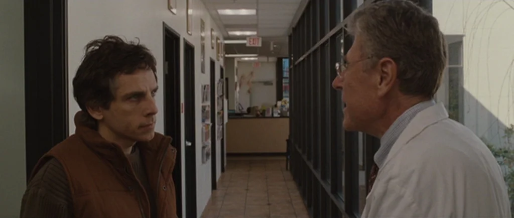 Greenberg (2010) movie still: medium — A sterile building hallway with white walls and a long bank of dark-framed windows. Two men face each other…
