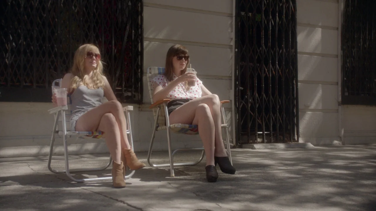 The Heart Machine (2014) movie still: fullbody — Two young women lounge on a concrete sidewalk in front of a building facade. They sit in portable…