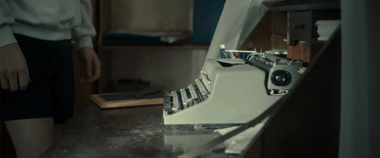 Jump, Darling (2022) movie still: medium — A worn wooden desk holds an off-white vintage manual typewriter in a dim room. A person stands partially…