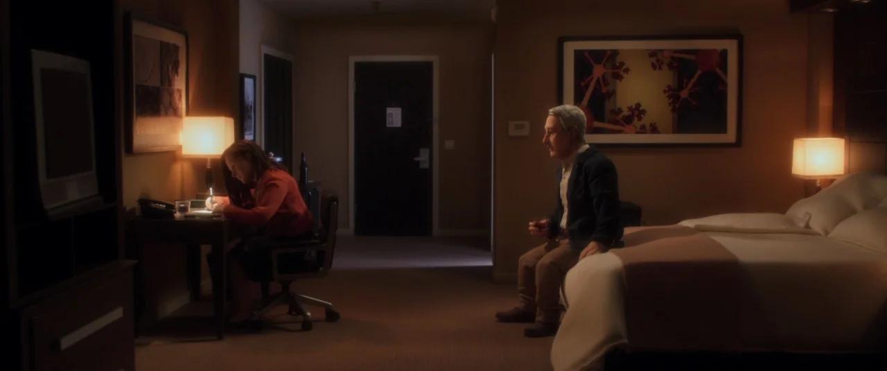 Anomalisa (2015) movie still: wide — A dimly lit hotel room features a desk with a lamp and a bed. An elderly man sits on the bed edge while a woman…